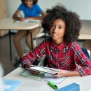 Happy Afro American schoolgirl looking at teacher holding tablet in classroom.